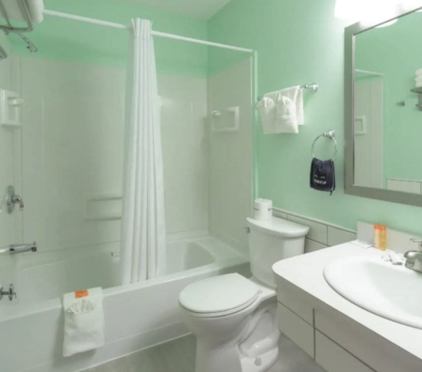 Light green bathroom with a bathtub, sink, and toilet, featuring white towels, a silver-framed mirror, and bright lighting above the vanity.