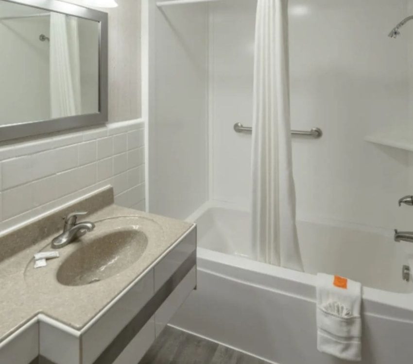 Simple bathroom with a beige sink countertop, silver-framed mirror, overhead lights, and white shower curtain over a bathtub with a grab bar.