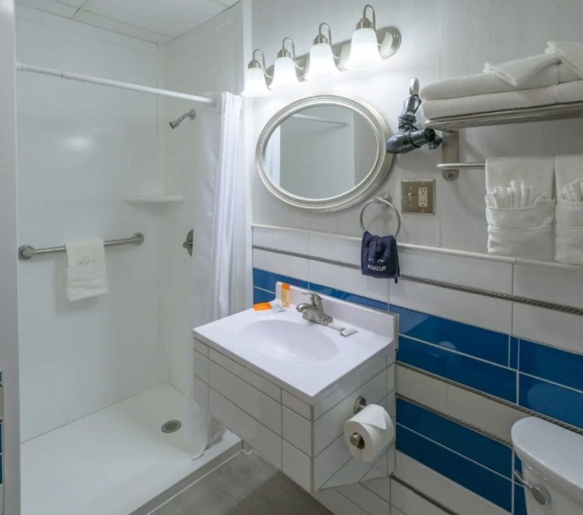 Neat and modern bathroom with a shower, sink, and oval mirror, featuring blue and white tile walls, folded towels, and a wall-mounted hairdryer.