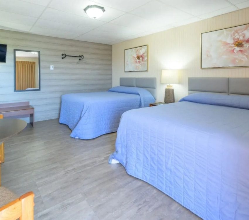Two queen beds with light blue bedding in a cozy, well-lit motel room featuring floral artwork, wood floors, and a small seating area.