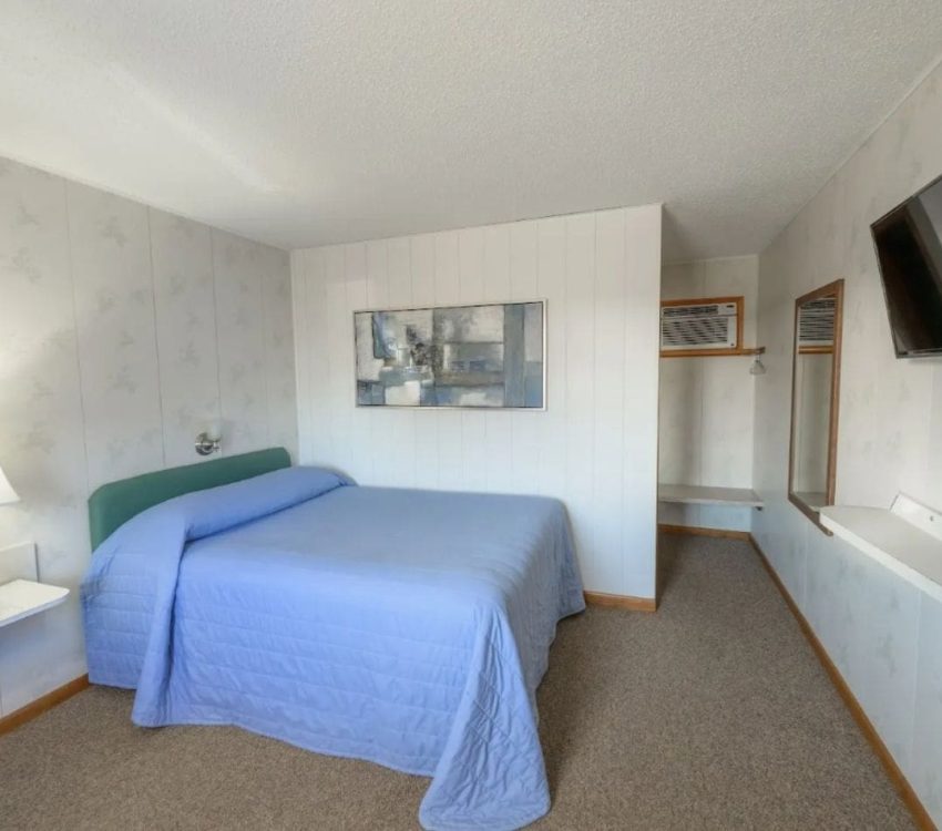 Queen bed with blue quilt and green headboard in a tidy motel room featuring a wall-mounted TV, abstract artwork, open closet, and carpeted floor.